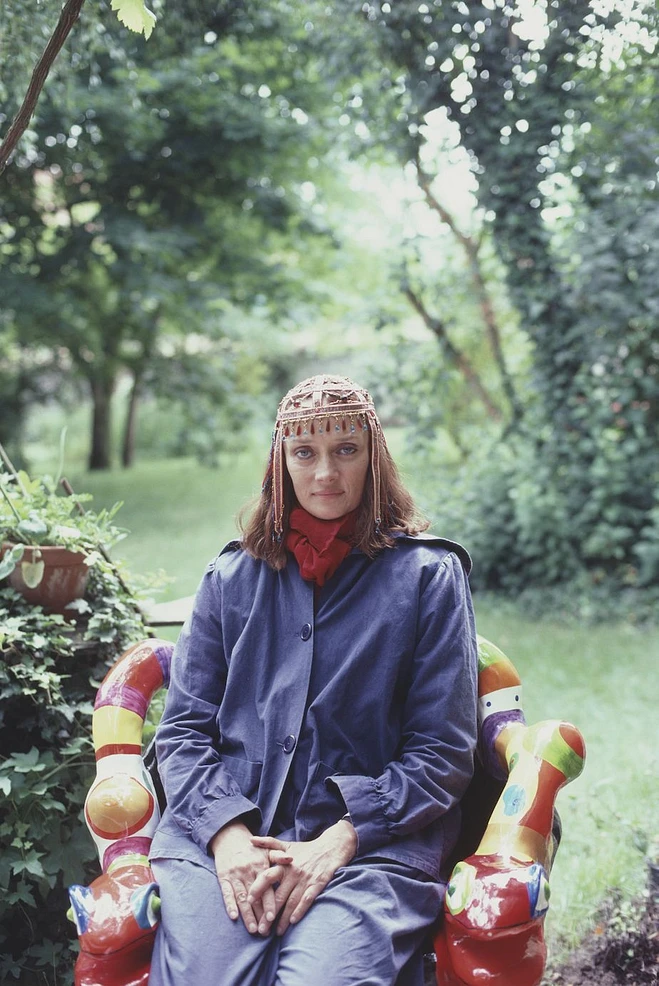 L'artiste Niki de Saint Phalle assise sur une chaise sculpturale, dans un jardin.